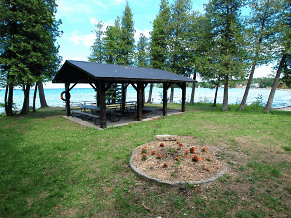 The small pavilion at Hibou Conservation Area with grass in the foreground, cedar trees behind it, and the lake in the background.